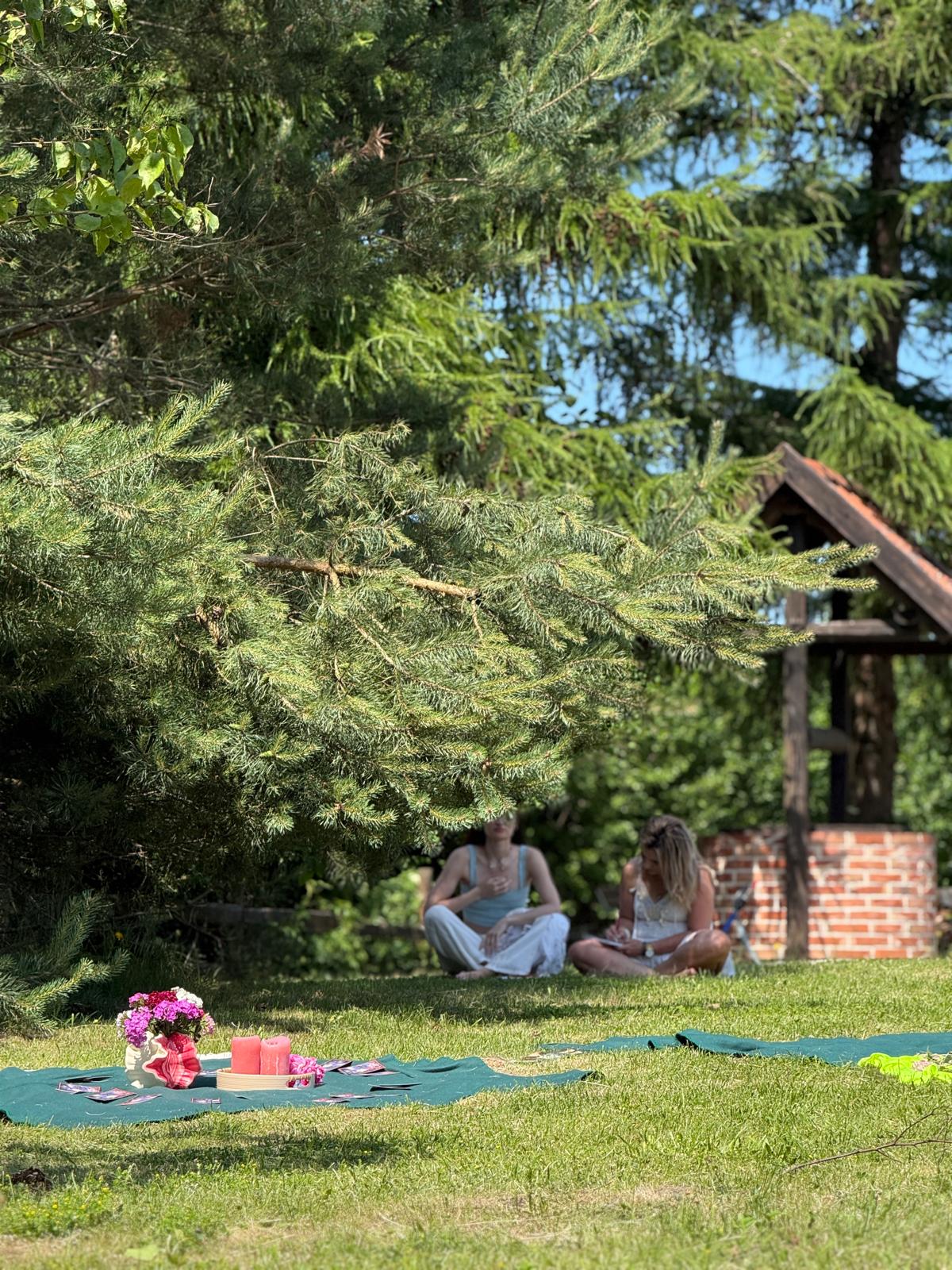 Dwie kobiety siedzą na trawie pod sosną obok kocu ze świecami i kwiatami