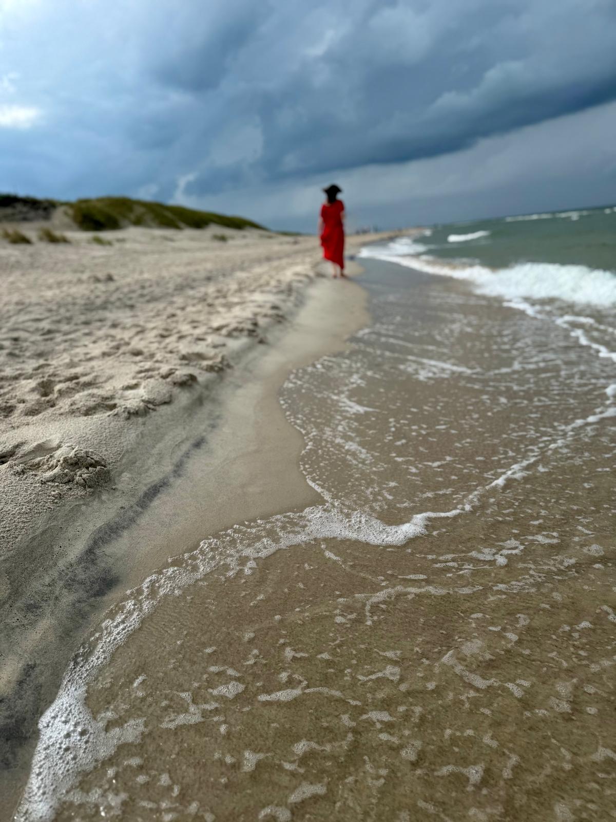 Kobieta w czerwonej sukni idzie wzdłuż linii wody na plaży pod ciemnym niebem